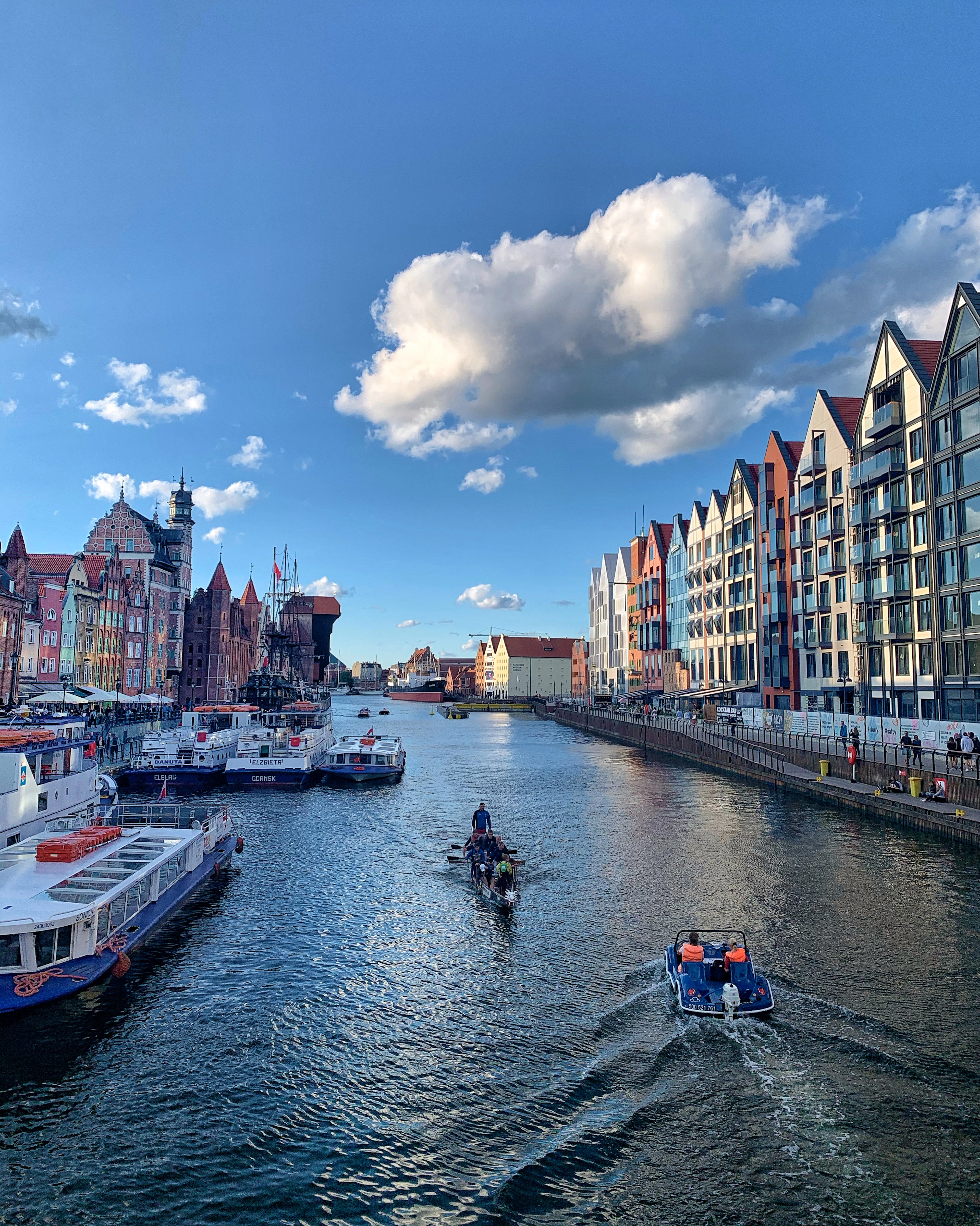 An elevated view over Gdańsk's Motława River, with tour boats, a kayaker and a motorboat passing colorful waterfront townhouses.