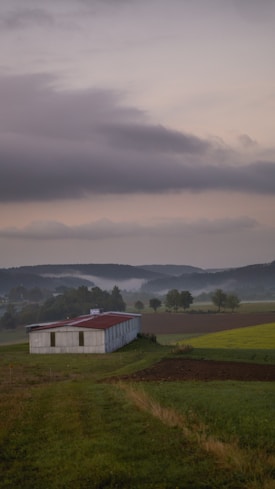 A rustic barn sits amidst a tranquil rural landscape with lush green fields and rolling hills in the background under an overcast sky.
