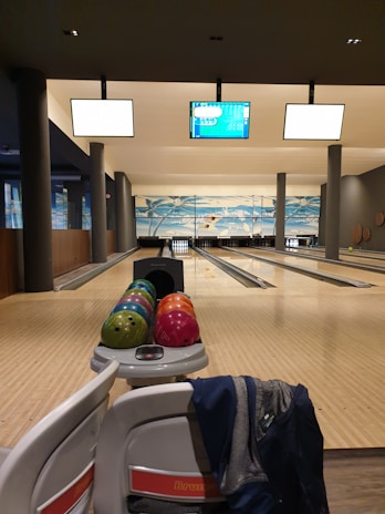 A bowling alley with several lanes and a rack of colorful bowling balls ready for play. Overhead screens display information for the games. The setting is well-lit and clean, with seating areas for players.