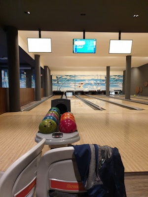 A bowling alley with several lanes and a rack of colorful bowling balls ready for play. Overhead screens display information for the games. The setting is well-lit and clean, with seating areas for players.