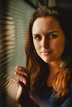 A person with long brown hair and blue eyes stands next to a textured glass window, softly lit from the side, creating gentle shadows. The expression is calm and contemplative, with a slight smile. The ambient light casts a warm glow on the hair and face.