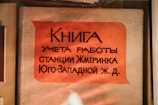 A book cover with text in Cyrillic script printed on it, featuring a large orange label with black lettering. The background is a neutral fabric, and the setting suggests it might be in a display or archive setting, possibly behind glass.