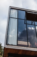 A modern architectural design featuring a large glass window with a wooden frame. The interior is partially visible, showing light curtains and a wooden platform or railing. The window reflects a cloudy sky, adding an open and airy feel to the structure.