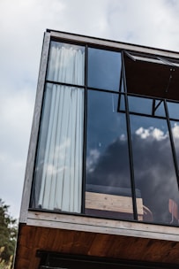 A modern architectural design featuring a large glass window with a wooden frame. The interior is partially visible, showing light curtains and a wooden platform or railing. The window reflects a cloudy sky, adding an open and airy feel to the structure.