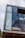 A modern architectural design featuring a large glass window with a wooden frame. The interior is partially visible, showing light curtains and a wooden platform or railing. The window reflects a cloudy sky, adding an open and airy feel to the structure.