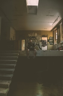 A dimly lit, abandoned space features graffiti-covered walls. A person sits on the edge of an elevated platform, partially illuminated by light coming through a small window. The environment exudes a sense of neglect and urban decay.
