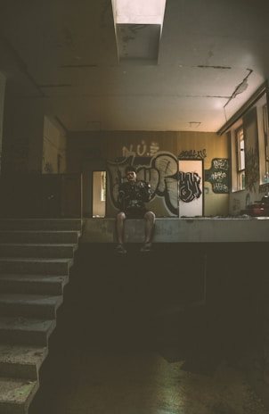 A dimly lit, abandoned space features graffiti-covered walls. A person sits on the edge of an elevated platform, partially illuminated by light coming through a small window. The environment exudes a sense of neglect and urban decay.