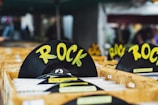A collection of vinyl records is organized in wooden crates, prominently featuring a black disc labeled with the genre 'ROCK' in bold yellow letters. Other records in the crates include artists such as Paul McCartney and The Beatles. The setting appears to be a market or store with a blurred background.