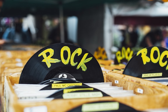 A collection of vinyl records is organized in wooden crates, prominently featuring a black disc labeled with the genre 'ROCK' in bold yellow letters. Other records in the crates include artists such as Paul McCartney and The Beatles. The setting appears to be a market or store with a blurred background.