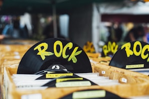 A collection of vinyl records is organized in wooden crates, prominently featuring a black disc labeled with the genre 'ROCK' in bold yellow letters. Other records in the crates include artists such as Paul McCartney and The Beatles. The setting appears to be a market or store with a blurred background.