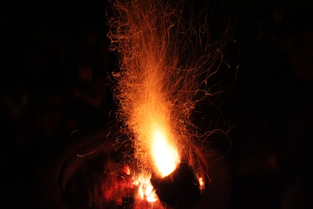 Intense flames and sparks rise dramatically from a fire, casting a warm glow. The black background accentuates the bright, fiery colors and the energy of the fire.