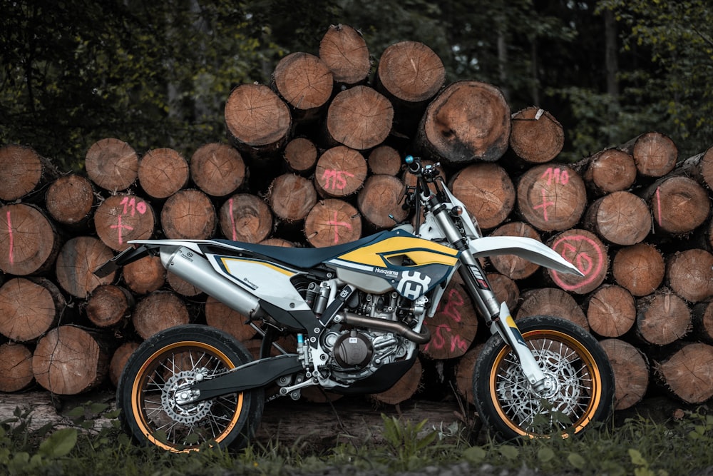 moto de cross de motocross negra, blanca y amarilla apoyada en una pila de troncos de árboles