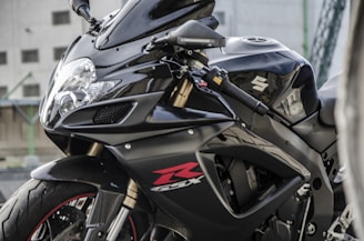 A sleek black motorcycle is parked, featuring sharp aerodynamic lines and a prominent Suzuki logo on the fuel tank. The design includes a glossy finish with red accents, noticeable on the fairing and tire sidewalls. The background shows a blurred urban setting with some concrete structures.