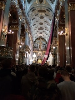 A large group of people gathered inside an ornate church. The ceiling is intricately decorated with gold accents and detailed artwork. The focal point appears to be towards the altar, which is equally elaborate with gold detailing and a prominent religious figure. There are chandeliers and columns on either side, contributing to the grandeur of the setting. A long, red fabric drapes down from the ceiling towards the altar.
