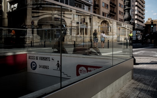An urban street scene with reflective glass showcasing pedestrians, city buildings, and a sign in a foreign language, suggesting a parking facility that operates 24 hours. The glass creates interesting visual reflections of the cityscape and people walking by.
