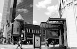 An urban street scene with a mix of architectural styles, featuring a modern glass skyscraper and an older brick building with a turret. Pedestrians are walking on the sidewalk, and the street is filled with activity.
