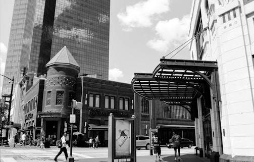 An urban street scene with a mix of architectural styles, featuring a modern glass skyscraper and an older brick building with a turret. Pedestrians are walking on the sidewalk, and the street is filled with activity.