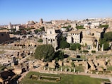A panoramic view of ancient Roman ruins with various historic structures, including columns, arches, and temples. Surrounding these ruins are lush green trees and vegetation. The background features a cityscape with buildings from different architectural periods against a clear blue sky.