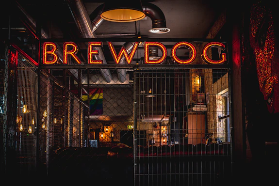 A dimly lit interior of a bar featuring a prominent neon sign that reads 'BREWDOG' in orange-red hues. The setting has industrial elements with visible metal pipes and fences, alongside warm wooden accents. A rainbow flag can be seen hanging in the background, and there are shelves with various items, possibly bottles or books, creating a cozy yet vibrant atmosphere.