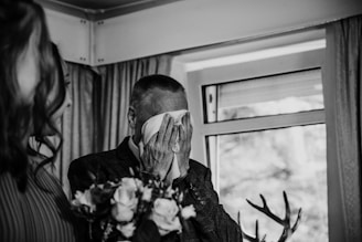 An elderly man is holding a handkerchief to his face, as he appears emotional, possibly wiping tears. A woman is partially visible on the left side, holding a bouquet of flowers. The setting is indoors with a window in the background, decorated with curtains.