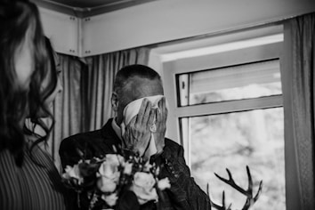 An elderly man is holding a handkerchief to his face, as he appears emotional, possibly wiping tears. A woman is partially visible on the left side, holding a bouquet of flowers. The setting is indoors with a window in the background, decorated with curtains.