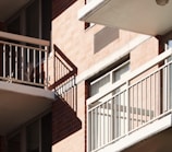 Balconies with metal railings casting distinct shadows on a brick facade of an apartment building. Sunlight illuminates the scene, highlighting the architectural features and creating a geometric pattern with the shadows.