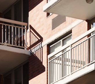 Balconies with metal railings casting distinct shadows on a brick facade of an apartment building. Sunlight illuminates the scene, highlighting the architectural features and creating a geometric pattern with the shadows.