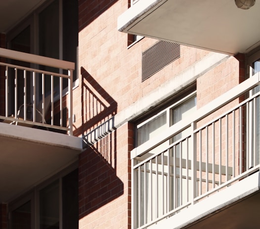 Balconies with metal railings casting distinct shadows on a brick facade of an apartment building. Sunlight illuminates the scene, highlighting the architectural features and creating a geometric pattern with the shadows.