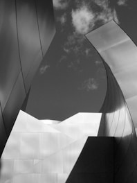 A black and white photograph capturing abstract architectural elements with curved and angular metal panels. The composition incorporates a cloudy sky in the background, creating a dynamic interplay of light and shadow across the surfaces.