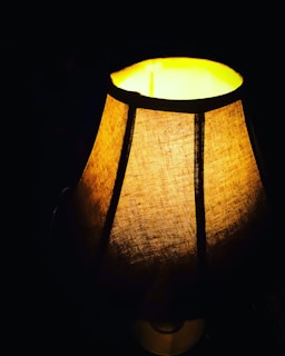 A warmly lit fabric lampshade creates a soft, cozy glow against a dark background. The light enhances the textured pattern of the fabric, contributing to an intimate and inviting ambiance.