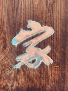 A wooden surface featuring a carved artistic symbol with a combination of light blue and beige colors. The background shows detailed wood grain textures, and there is a small stamp or seal in the bottom right corner.