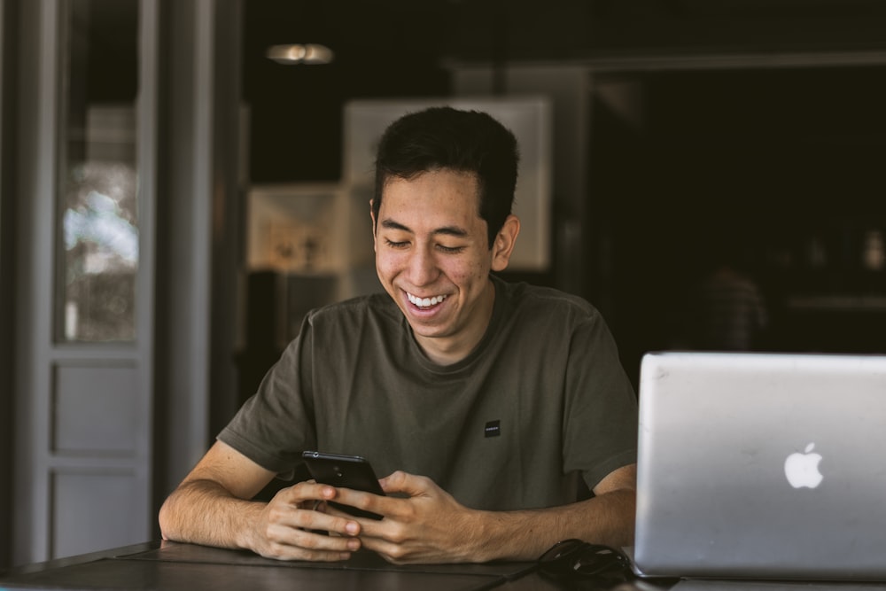 close-up photography of smiling man while holding phone beside silver MacBook