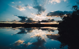 The image captures a serene landscape with a stunning sunset reflected perfectly on the surface of a calm lake. Clouds scattered across the sky mirror beautifully on the water, creating a symmetrical and tranquil scene. Silhouettes of trees frame the water, adding depth and contrast to the vibrant sky.
