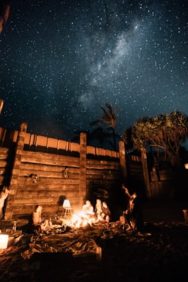 A group of people are gathered around a campfire on a sandy area, surrounded by a wooden fence and a few trees under a clear, starry night sky with the Milky Way visible. Some people are seated, and the warm glow of the fire illuminates their figures, creating a cozy and relaxed atmosphere.