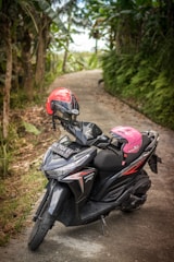 A black scooter is parked on a narrow, winding pathway surrounded by lush greenery. The scooter has two helmets resting on it, one red and one pink. The background features dense foliage and tropical plants.