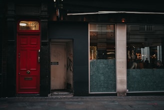 A narrow urban street scene features a striking red door with a windowpane that displays the address 36-38 Glasshouse St. Adjacent to the door is a small entrance labeled as a fire exit, marked by graffiti. Large glass windows with patterned designs suggest a modern interior space, where a few people can be seen seated indoors.