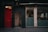 A narrow urban street scene features a striking red door with a windowpane that displays the address 36-38 Glasshouse St. Adjacent to the door is a small entrance labeled as a fire exit, marked by graffiti. Large glass windows with patterned designs suggest a modern interior space, where a few people can be seen seated indoors.