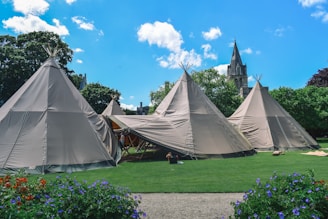 A cluster of large canvas teepees is set up on a well-manicured lawn. The scene includes vibrant green grass and colorful flowers in the foreground, creating a contrast with the beige of the tents. A gothic-style church with a tall spire is visible in the background, partially obscured by lush green trees under a bright blue sky with a few fluffy clouds.