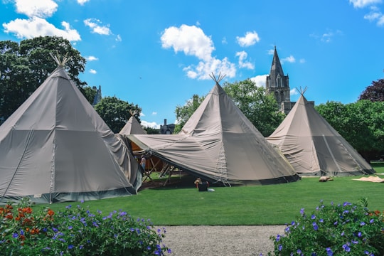 A cluster of large canvas teepees is set up on a well-manicured lawn. The scene includes vibrant green grass and colorful flowers in the foreground, creating a contrast with the beige of the tents. A gothic-style church with a tall spire is visible in the background, partially obscured by lush green trees under a bright blue sky with a few fluffy clouds.