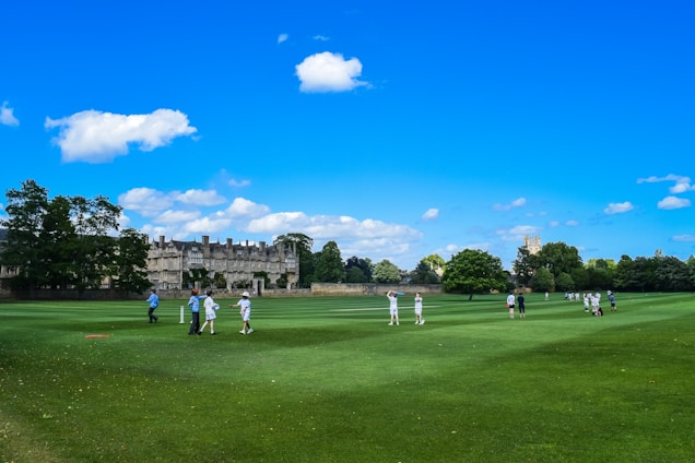 A group of people playing cricket on a large, well-maintained green field on a sunny day. The field is surrounded by a historic building and lush green trees. The clear blue sky is dotted with a few fluffy white clouds, adding to the serene atmosphere.