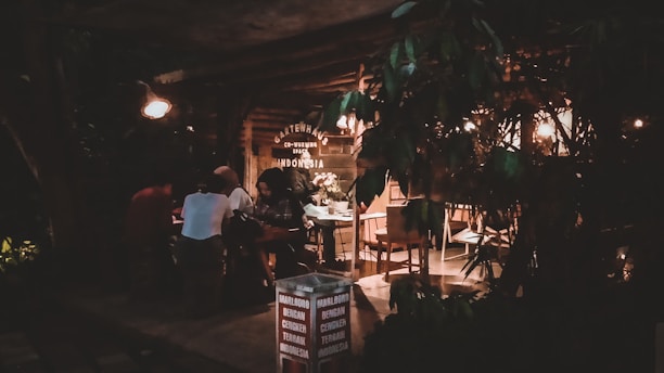A dimly lit cafe or coworking space with several people sitting at a table. The environment appears cozy with warm lighting, wooden furniture, and foliage partially blocking the view. Signage for Marlboro cigarettes and a location, Canggu, Indonesia, is visible.