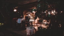 A dimly lit cafe or coworking space with several people sitting at a table. The environment appears cozy with warm lighting, wooden furniture, and foliage partially blocking the view. Signage for Marlboro cigarettes and a location, Canggu, Indonesia, is visible.