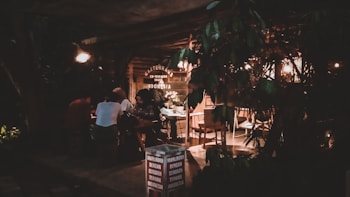 A dimly lit cafe or coworking space with several people sitting at a table. The environment appears cozy with warm lighting, wooden furniture, and foliage partially blocking the view. Signage for Marlboro cigarettes and a location, Canggu, Indonesia, is visible.