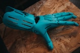 A robotic hand in a bright teal color lies on a textured wooden surface. The design of the hand includes articulated joints and slits on each finger, suggesting functionality and mechanical sophistication.