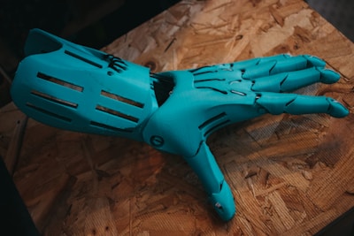 A robotic hand in a bright teal color lies on a textured wooden surface. The design of the hand includes articulated joints and slits on each finger, suggesting functionality and mechanical sophistication.