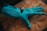 A robotic hand in a bright teal color lies on a textured wooden surface. The design of the hand includes articulated joints and slits on each finger, suggesting functionality and mechanical sophistication.