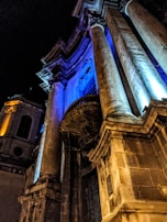 A dramatic architectural facade illuminated by vibrant blue and amber lights, highlighting tall stone columns and intricate structural details against the night sky.
