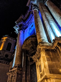 A dramatic architectural facade illuminated by vibrant blue and amber lights, highlighting tall stone columns and intricate structural details against the night sky.