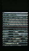 A window with metal grillwork featuring geometric patterns provides a view of the outside. Beyond the grill, layers of horizontal slats, possibly blinds or louvered panels, partially obscure the view of a garden with green foliage and some tiled roofs in the background.