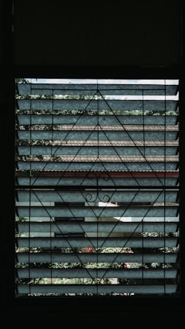 A window with metal grillwork featuring geometric patterns provides a view of the outside. Beyond the grill, layers of horizontal slats, possibly blinds or louvered panels, partially obscure the view of a garden with green foliage and some tiled roofs in the background.